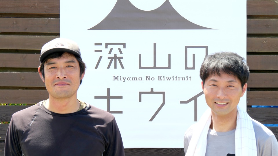 深山のキウイの看板の前に立つ深井さん（左）と山田さん（右）